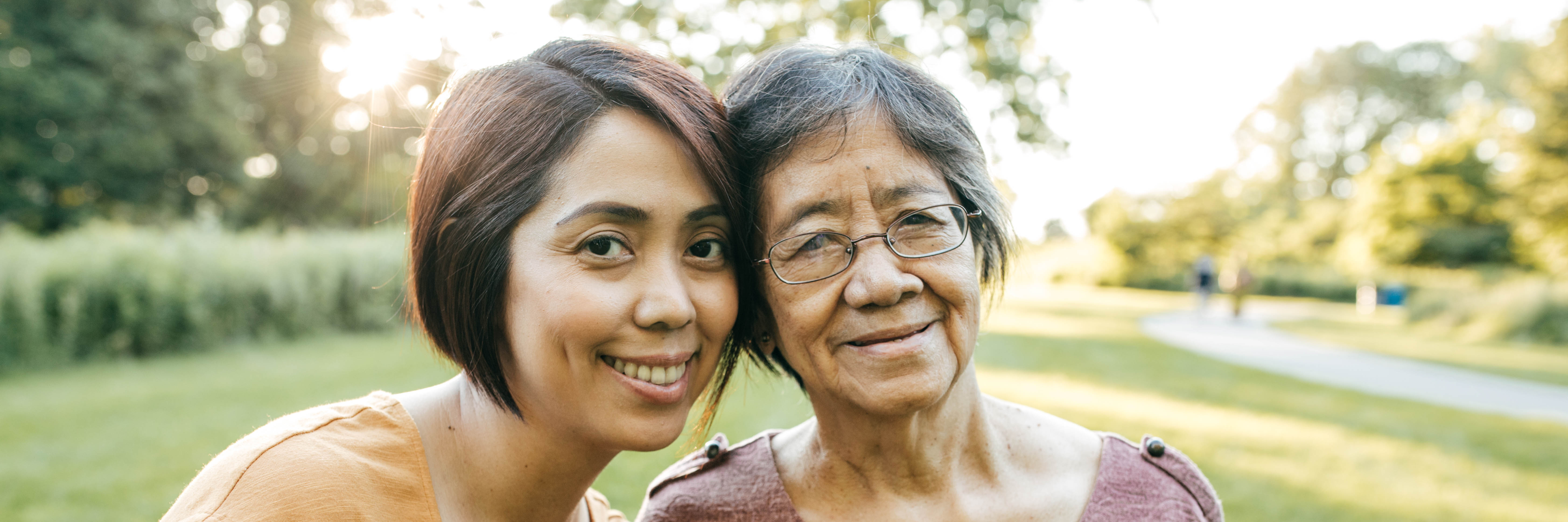 Middle-aged woman standing close to her senior mother, with their heads touching and smiling, outdoors on a sunny day at a park.
